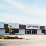 A "glo tanning" storefront with pink neon letters, a white and gray facade, large windows, and a paved parking lot.