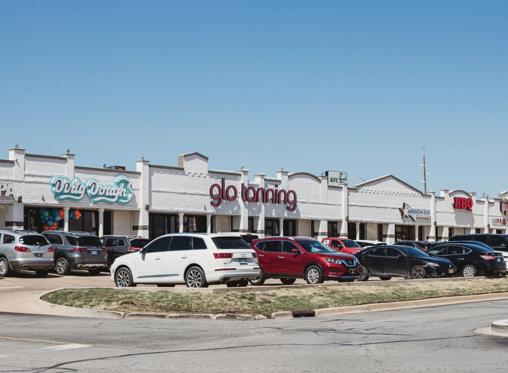 A row of storefronts with white brick facades, including "Dirty Dough," "glo tanning," "KFC," and "BBQ," with parked cars in front.