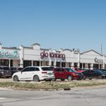 A row of storefronts with white brick facades, including "Dirty Dough," "glo tanning," "KFC," and "BBQ," with parked cars in front.