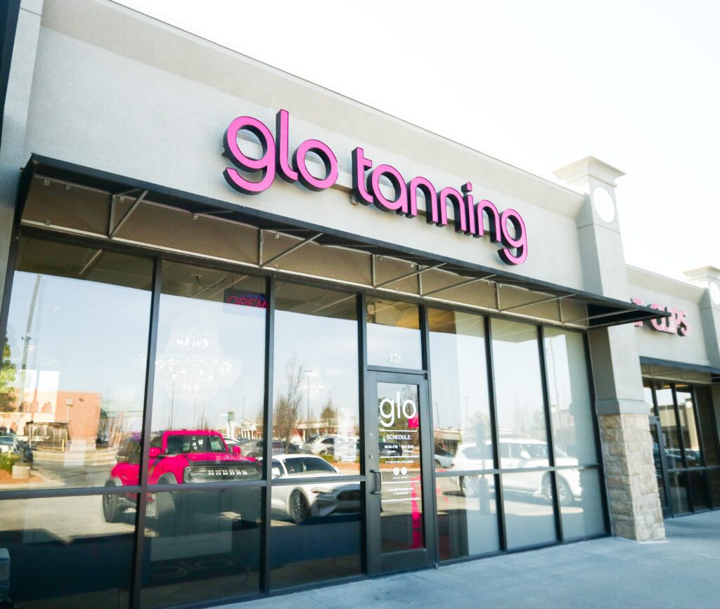 A Glo Tanning storefront with pink neon letters, a glass door, and large windows, reflecting a white car and a red pickup truck.