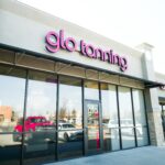 A Glo Tanning storefront with pink neon letters, a glass door, and large windows, reflecting a white car and a red pickup truck.