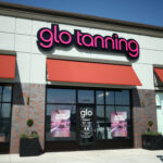 A Glo Tanning storefront with pink neon letters, red awnings, glass doors with "Now Open" signs, and potted plants.