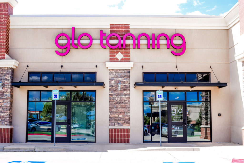 A Glo Tanning storefront with pink neon letters, two glass doors, and a stone facade, under a partly cloudy sky.