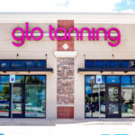 A Glo Tanning storefront with pink neon letters, two glass doors, and a stone facade, under a partly cloudy sky.