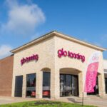 A Glo Tanning storefront with pink illuminated signage on a light brick building under a blue sky. A "NOW OPEN!" flag waves.