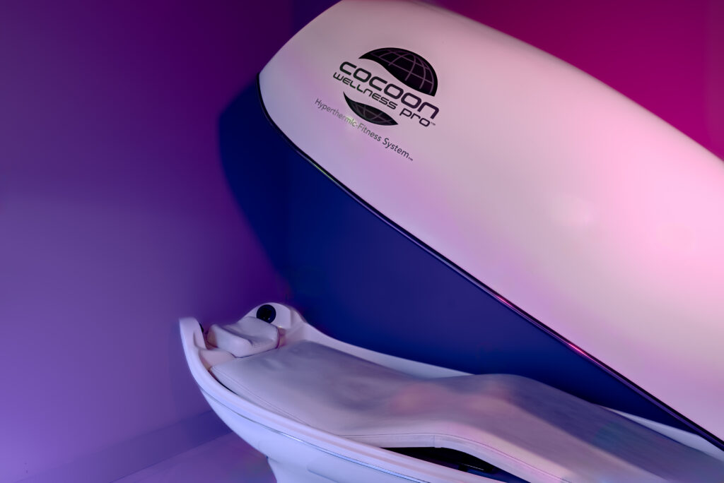 A "Cocoon Wellness Pro" pod with the text "Hyperthermic Fitness System." The wall behind is illuminated by purple light,