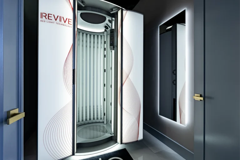 A standing red light therapy booth with "REVIVE" and "RED LIGHT TECHNOLOGY" text, a dark gray door and a dark mirror on the wall.