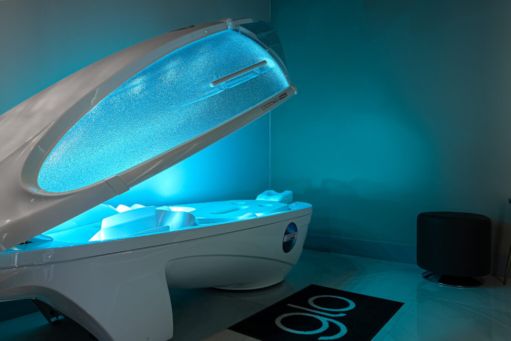 A white Skin Wellness Pro pod with a glowing blue interior, a black rug with "glo" text, and a dark stool in the corner.